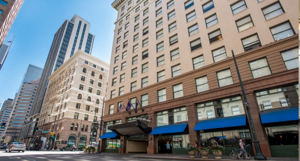The image shows a city street lined with tall buildings, including one with blue awnings and flags at the entrance, on a bright, sunny day.