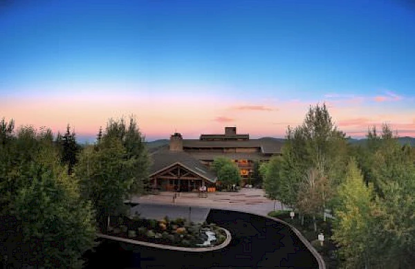 A picturesque lodge surrounded by trees during sunset, featuring a winding driveway, a rugged roofline, and a beautifully landscaped entrance.