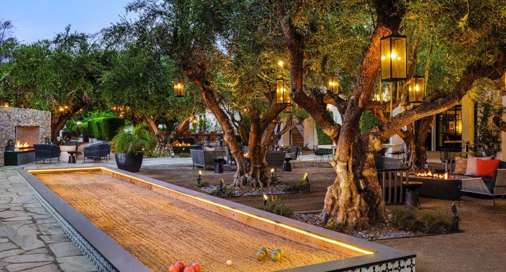 An outdoor area with a bocce ball court, surrounded by large trees, lanterns, and cushioned seating, creating a cozy, well-lit atmosphere.