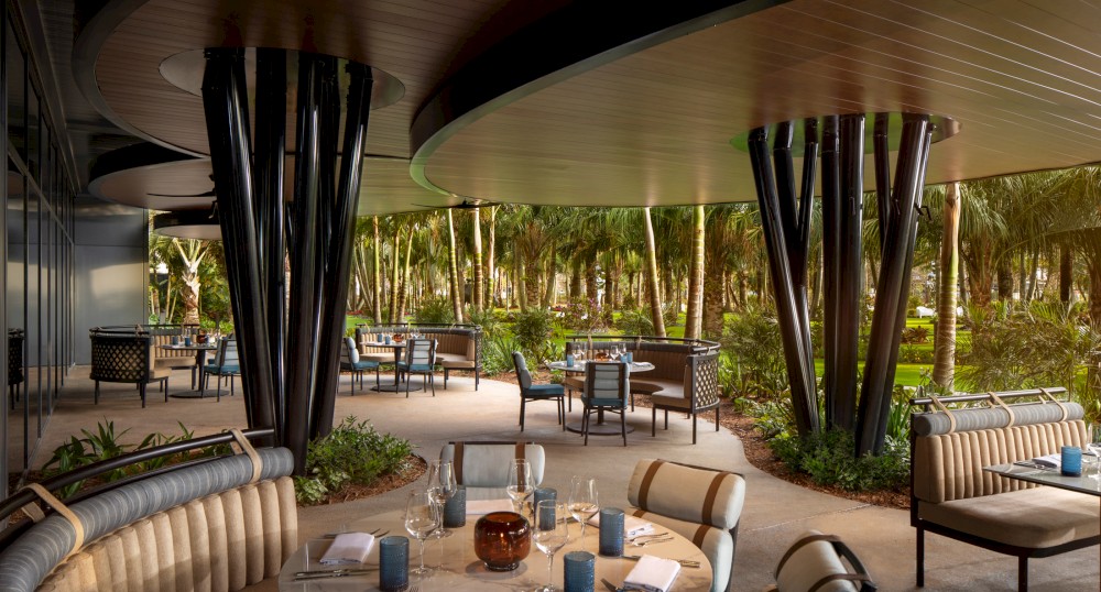 The image shows an outdoor dining area with elegant seating arrangements, modern architectural design, and a lush green garden backdrop.