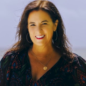 The image shows a woman with long dark hair wearing hoop earrings, a dark patterned top, and a necklace with a pendant, smiling at the camera.