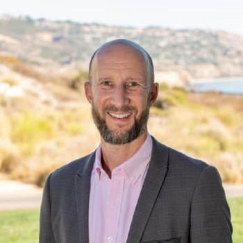 A man with a beard and a bald head is standing outdoors, wearing a grey suit jacket and a pink shirt, smiling at the camera.