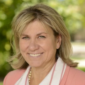 A woman with blonde hair is smiling, wearing a pink blazer and white shirt, standing outdoors with green foliage in the background.