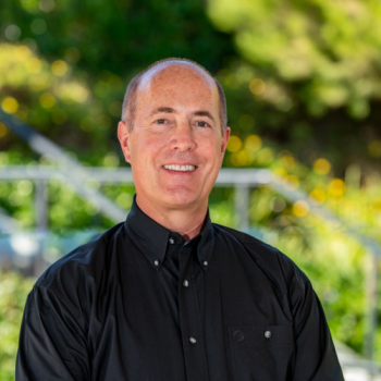 A person in a black shirt smiles at the camera, with a green, out-of-focus background.