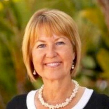 The image shows a smiling woman with short blonde hair, wearing a black top, white necklace, and pearl earrings, with a blurred green background.