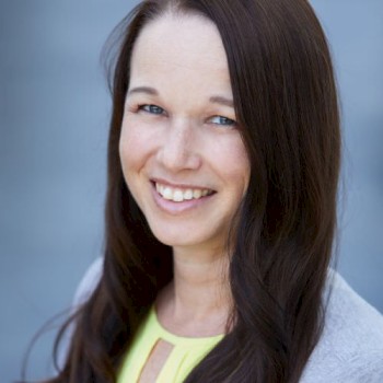 A person with long dark hair smiles at the camera, wearing a yellow top and a gray sweater. The background is blurred in blue tones.