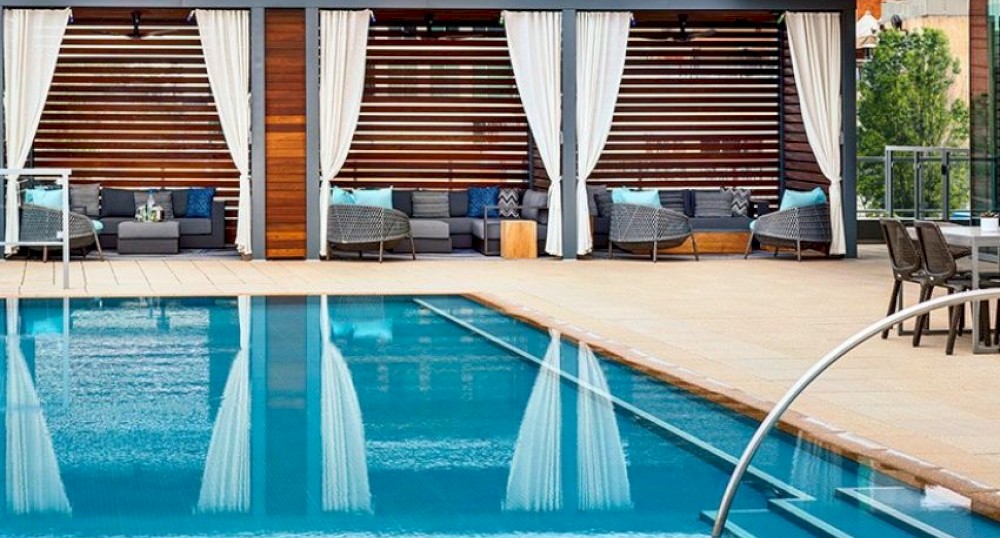 A luxurious outdoor pool area with lounge chairs, cabanas with curtains, and modern buildings in the background on a bright day.