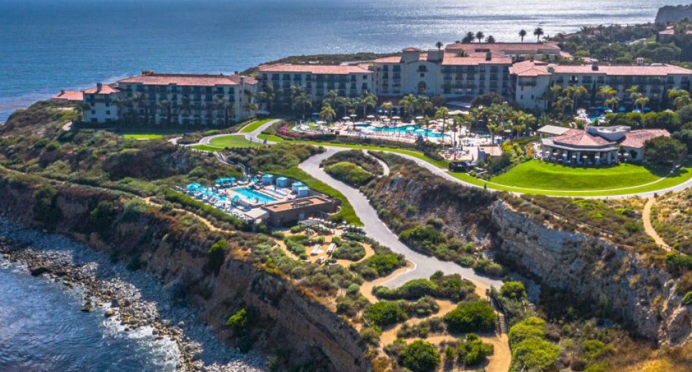 The image shows a coastal resort on a cliff with multiple buildings, swimming pools, and pathways, overlooking the ocean with a rocky shoreline.