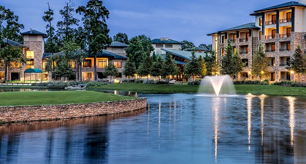 The image shows a serene scene of buildings by a tranquil lake with a fountain, surrounded by greenery, captured during twilight with lights reflecting off the water.