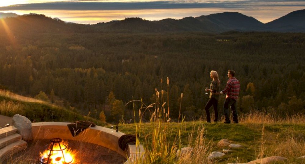 A couple standing near a fire pit on a grassy hill, overlooking a scenic forest and mountain landscape during sunset.