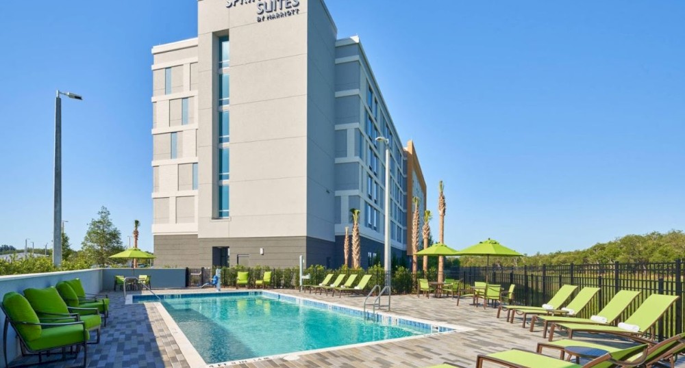 This image shows the exterior of a SpringHill Suites by Marriott hotel with a swimming pool, lounge chairs, and green umbrellas on a sunny day.