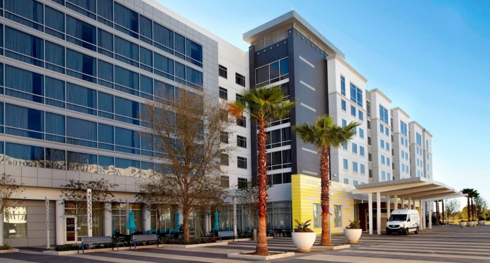 The image shows a modern multi-story building with large windows, palm trees, and a white van parked near the entrance.
