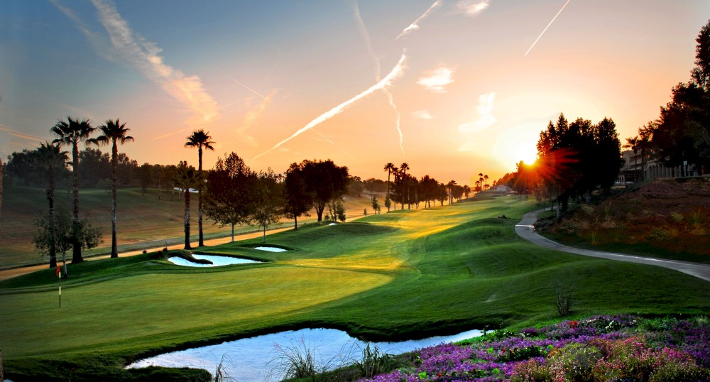 A scenic golf course is seen at sunset, featuring well-manicured greens, sand bunkers, water hazards, and vibrant flowers in the foreground.