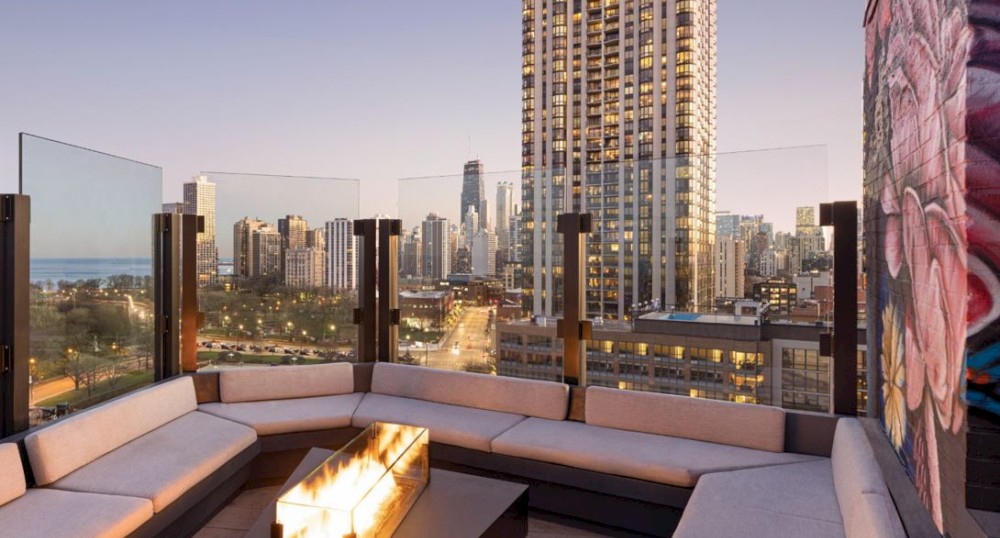 A rooftop lounge with a fire pit, surrounded by modern buildings and city skyline at dusk, with seating and a mural on the wall.
