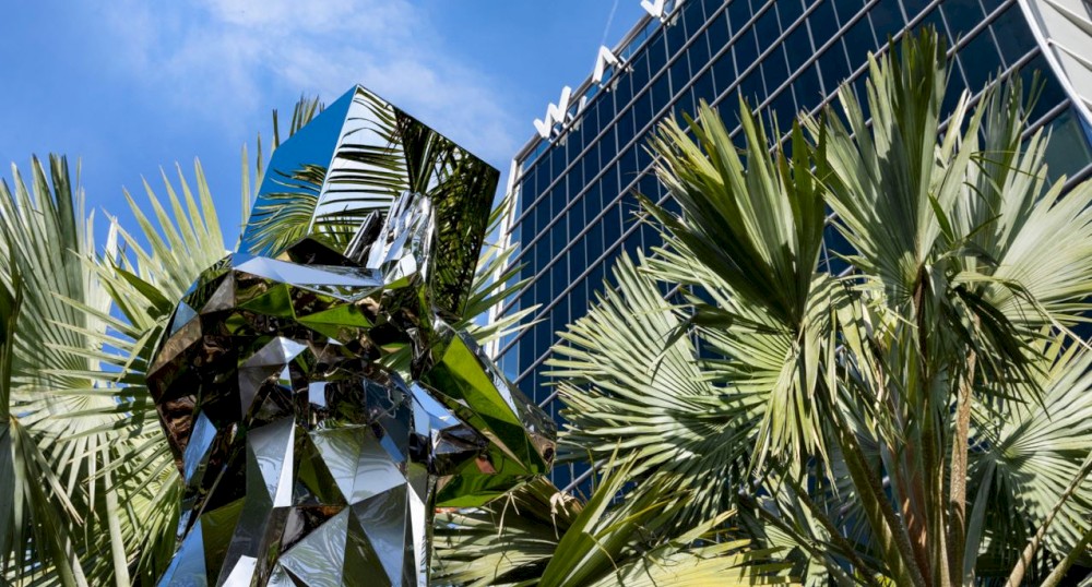 The image depicts a reflective, angular sculpture among palm trees with a modern glass building in the background against a blue sky.