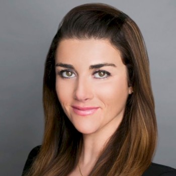 A woman with long brown hair is posing for a professional headshot against a grey background, wearing a black blazer and white top.