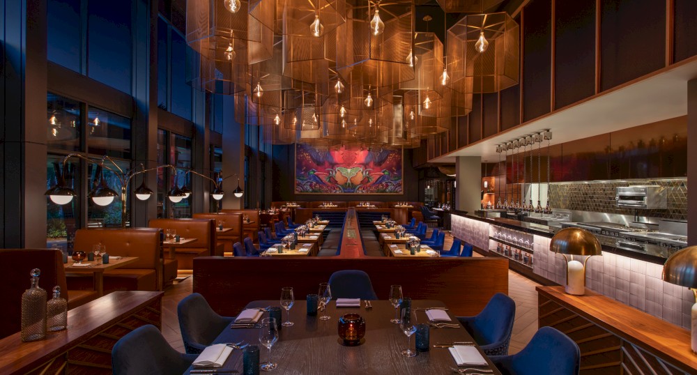 A modern, upscale restaurant with stylish lighting, wooden furnishings, and a mural on the back wall, featuring an open kitchen area and neatly set tables.