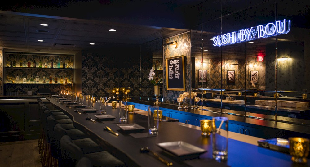 A dimly lit sushi bar with counter seating, featuring a neon sign "Sushi by Bou" and elegant decor with gold accents and framed art on the walls.
