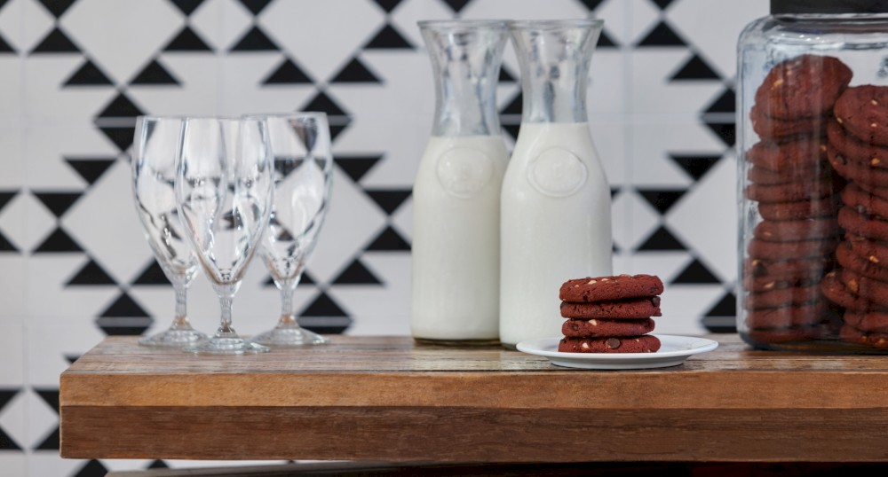 The image shows two carafes of milk, a stack of cookies on a plate, a large jar filled with cookies, and four empty glasses on a wooden surface.