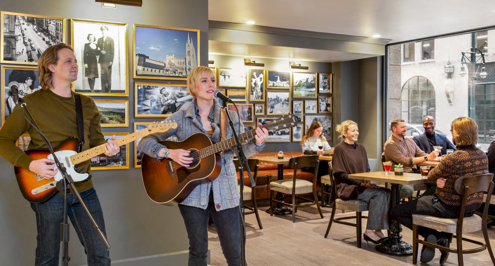 Musicians perform with guitars in a cozy cafe while patrons seated at tables enjoy their time and socialize, surrounded by framed pictures on the walls.