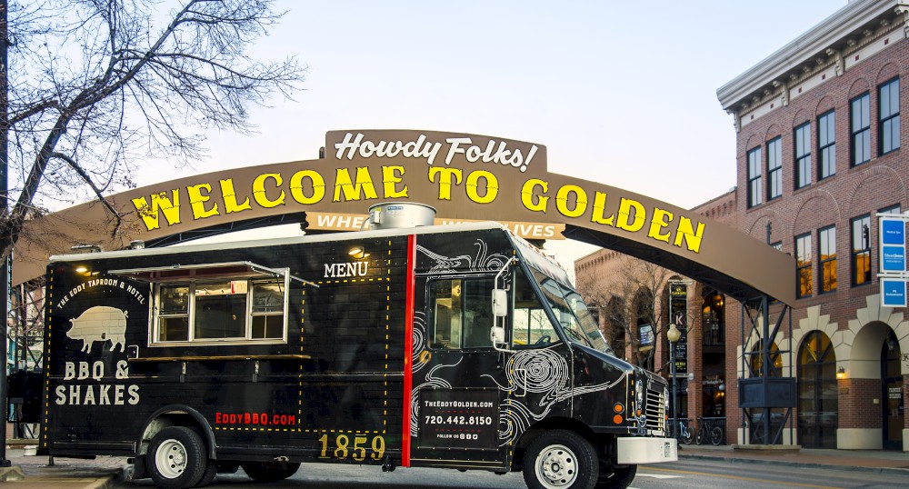 The image shows a black food truck labeled "BBQ & Shakes" parked under a "Welcome to Golden" sign in a street with historic buildings.