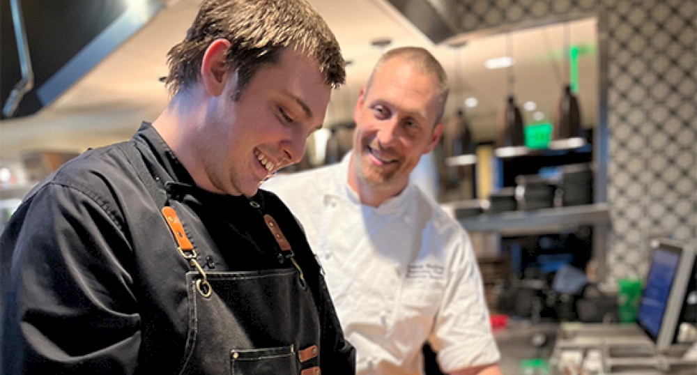 Two chefs in a kitchen, one in a black apron smiling and another in white observing, with kitchen equipment visible in the background.