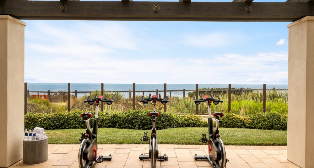 The image shows three stationary bikes on a patio overlooking a scenic outdoor view with greenery and the ocean in the background.