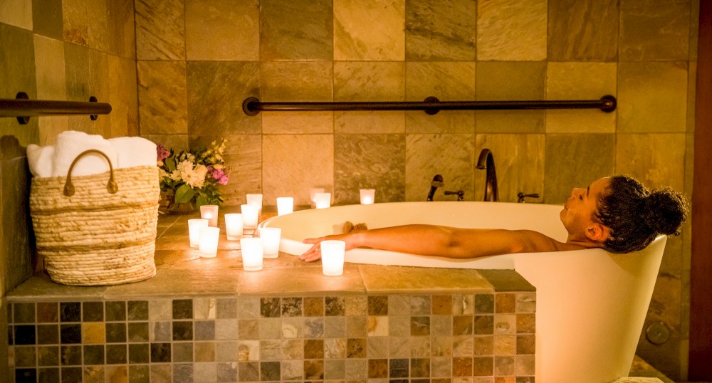 A person relaxes in a bathtub surrounded by lit candles, next to a basket with towels and flowers, in a beautifully tiled bathroom.