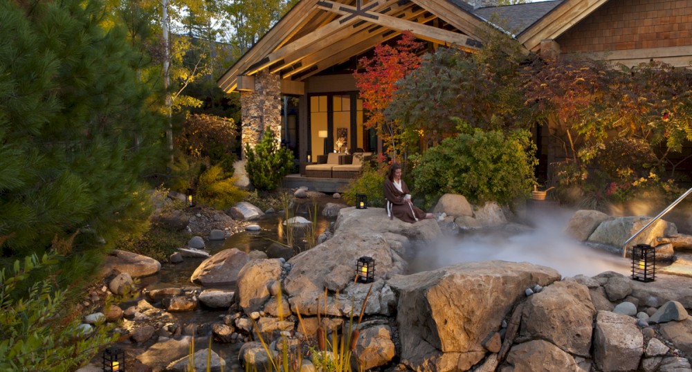 A rustic house surrounded by lush greenery, rocks, and a small pond is seen, with a woman sitting on a rock near lanterns.