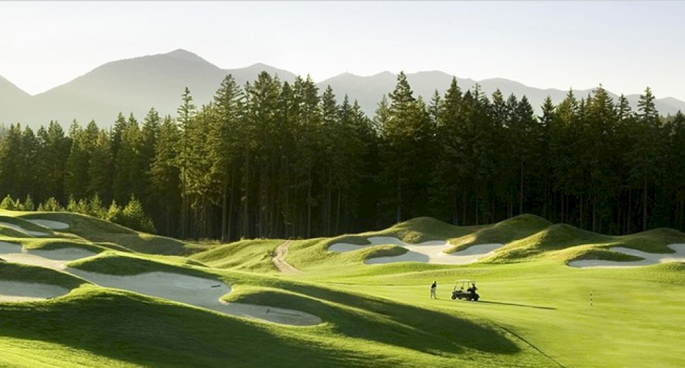 A scenic golf course with rolling green hills, sand bunkers, and a forested mountain backdrop. Two people are near a golf cart on the course.
