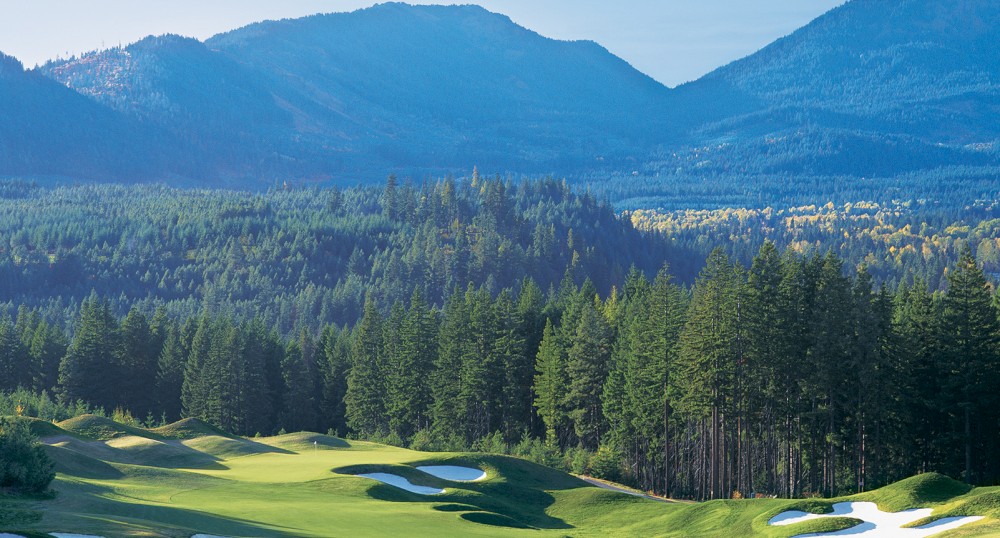 A scenic golf course with lush green fields, sand traps, and surrounded by dense forests against a backdrop of mountains under a clear blue sky.