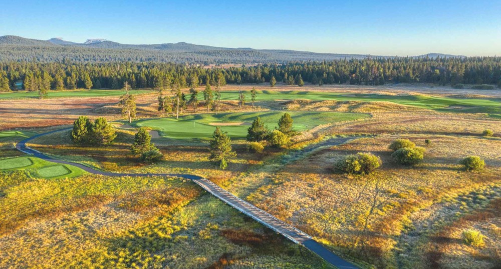 A scenic aerial view of a golf course with green fairways, trees, a small stream, and mountains in the background under a clear blue sky.