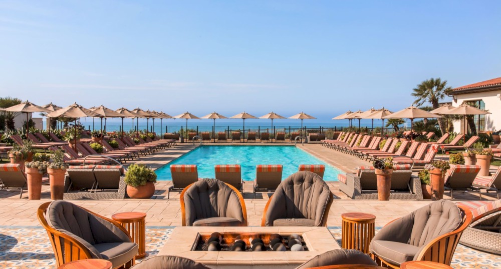 An outdoor seating area with a fire pit overlooks a pool surrounded by lounge chairs and umbrellas, with an ocean view in the background.