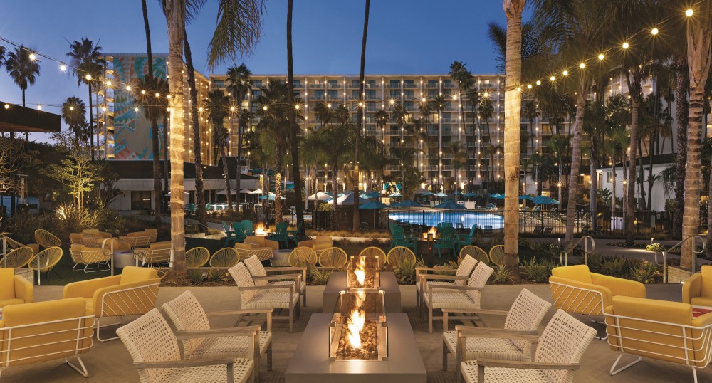 The image shows an outdoor seating area with fire pits, surrounded by palm trees and string lights, overlooking a pool and a large hotel at dusk.