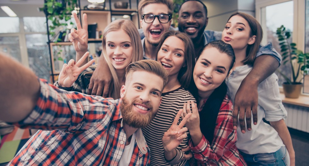 A group of seven smiling people taking a selfie in a bright room with large windows and plants in the background, many making peace signs.