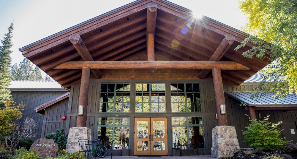 A building with a sloped roof, large wooden beams, and glass entrance doors, surrounded by trees and shrubs under a sunny sky.