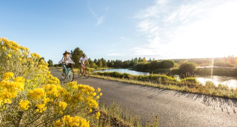 Two people cycling on a path beside a scenic river, surrounded by yellow flowers and greenery under a clear blue sky, in the late afternoon sunlight.