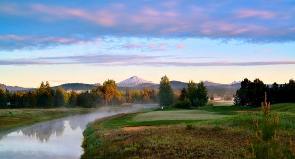 A peaceful landscape featuring a river, a green field, and a distant mountain under a partly cloudy sky during what appears to be sunrise.