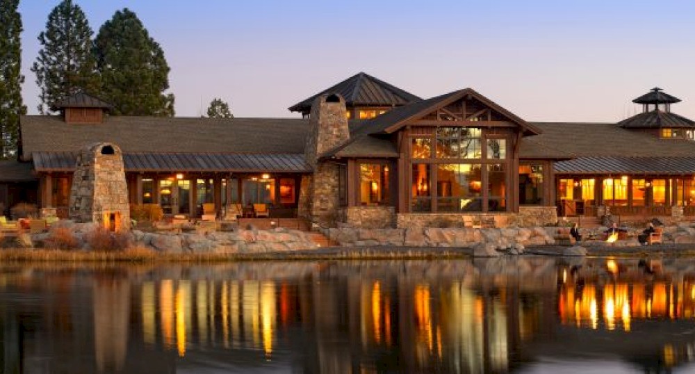 A large, rustic lodge-style building is reflected in a tranquil lake at sunset, with warm lights glowing through its numerous windows and trees in the background.