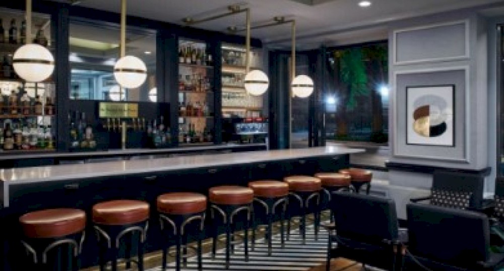 The image shows a modern bar with a clean design, featuring leather bar stools, contemporary lighting, and artwork on the wall, creating a stylish ambiance.