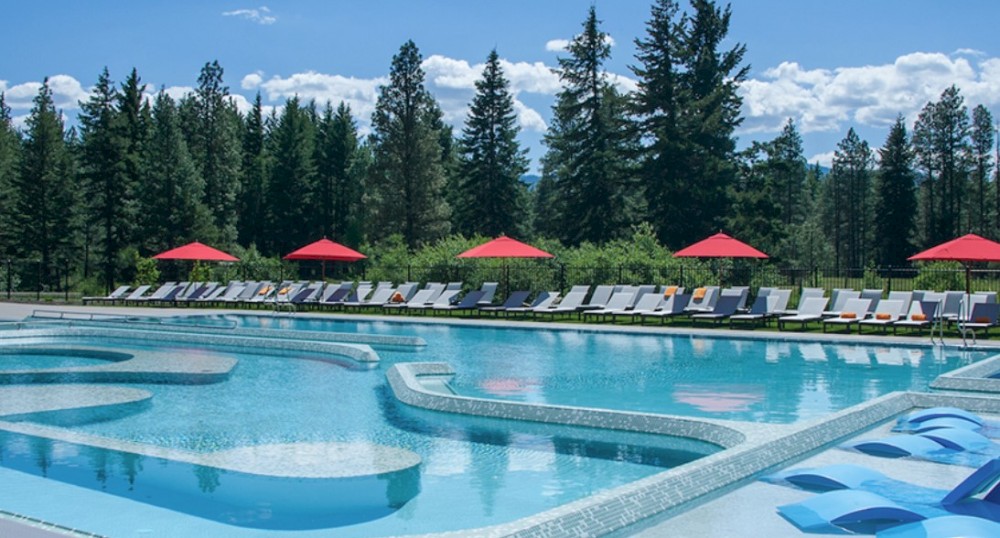 An outdoor pool area with sun loungers, red umbrellas, and a forested backdrop under a bright blue sky.