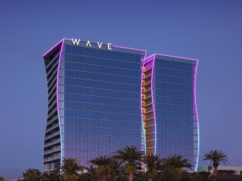 The image shows two modern, wave-shaped glass buildings with the name "WAVE" on top, illuminated with pink and purple lighting, with palm trees in the foreground.