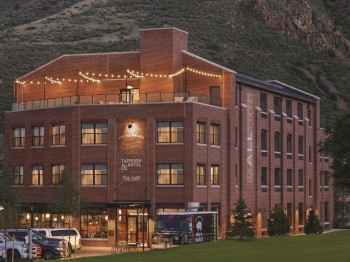 A multi-story brick building with lights on the top terrace, surrounded by cars and greenery, set against a backdrop of hills and trees.