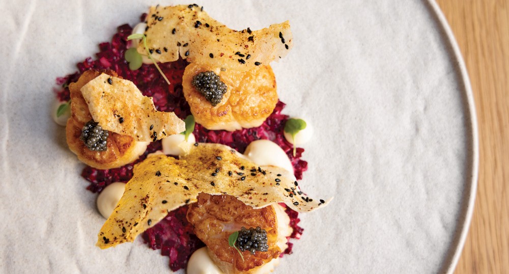 The image shows an elegant dish with seared scallops, crispy garnish, and caviar, set on a bed of beetroot and white sauce, on a white plate.