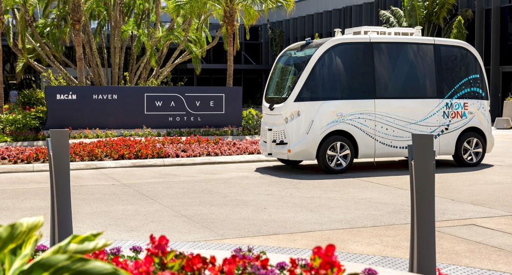 A small, white autonomous vehicle is parked in front of a building with a "Wave Hotel" sign among colorful landscaping.
