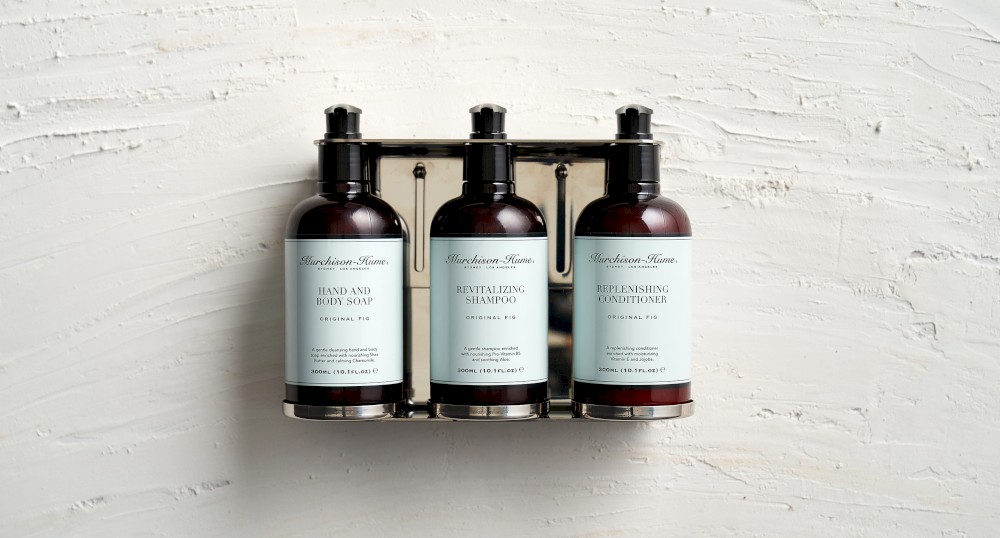 The image shows three bottles of hand and body care products mounted on a wall in a metal rack, likely shampoo, conditioner, and body wash.