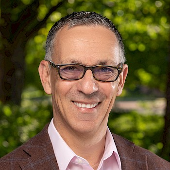 A smiling man wearing glasses, a suit jacket, and a pink shirt, standing outdoors with a green leafy background.
