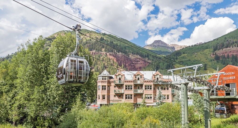 The image shows a gondola lift traveling over greenery with mountain views and buildings in the background under a partly cloudy sky.