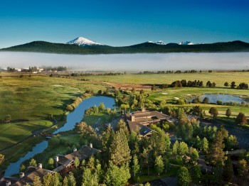 Scenic landscape with a river, trees, a village, and snow-capped mountains in the background under a clear blue sky, with mist in the distance.