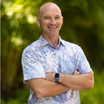A smiling person stands outdoors with arms crossed, wearing a floral shirt and a smartwatch, with lush greenery in the background.